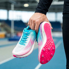 Person holding a pair of pink and blue running shoes with cleats on an outdoor track.