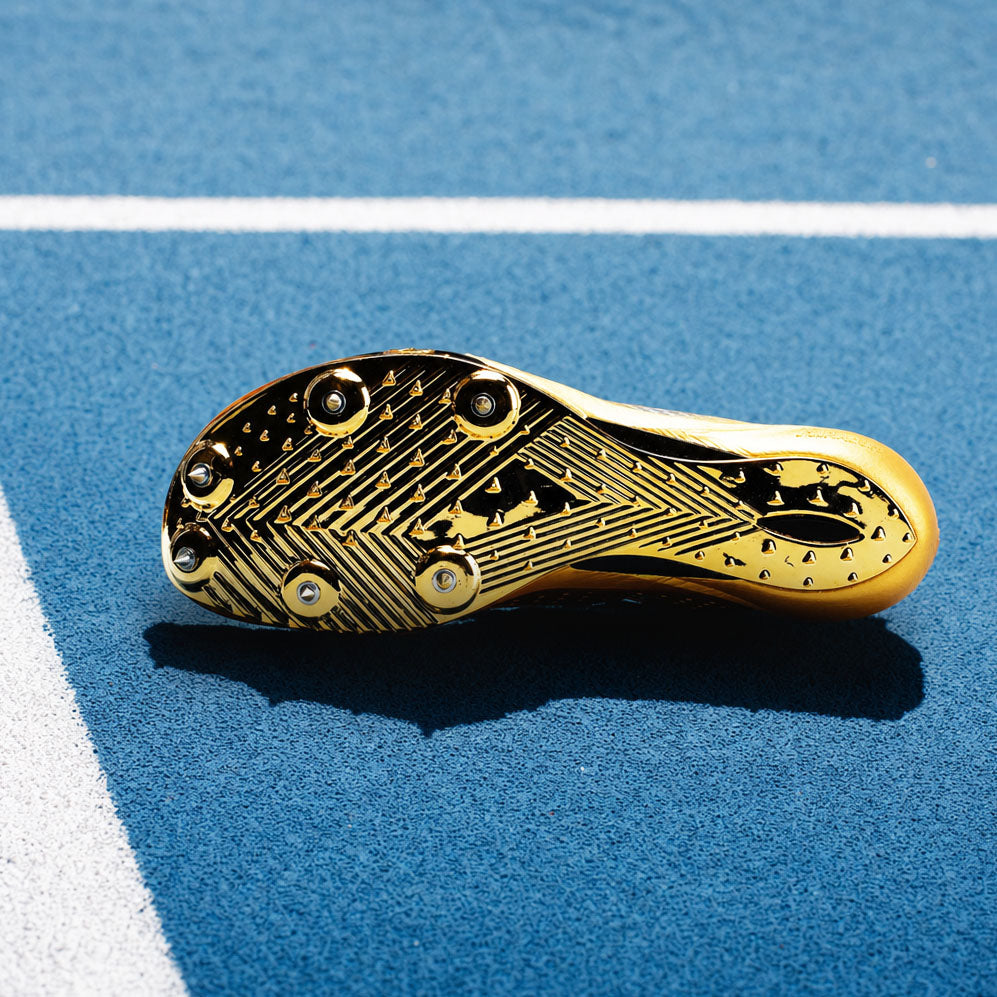 A close-up of a gold track cleat sole with black studs and tread patterns, resting on a blue running track with white lines.